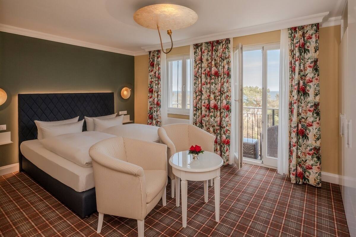 Ein Hotelzimmer mit einem großen Bett, zwei gestreiften Sesseln, einem kleinen runden Tisch mit Blumenvase und einem Balkon mit blumigen Vorhängen.