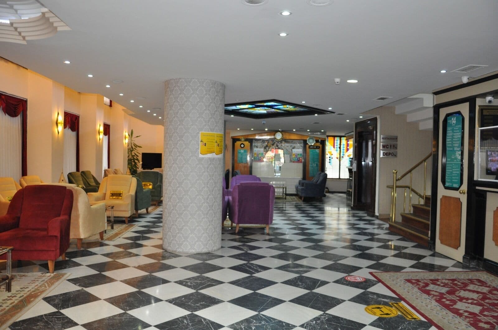 Un hall d’hôtel avec un sol à carreaux, des coins salon avec des fauteuils, un pilier central et un escalier menant aux toilettes (WC11).