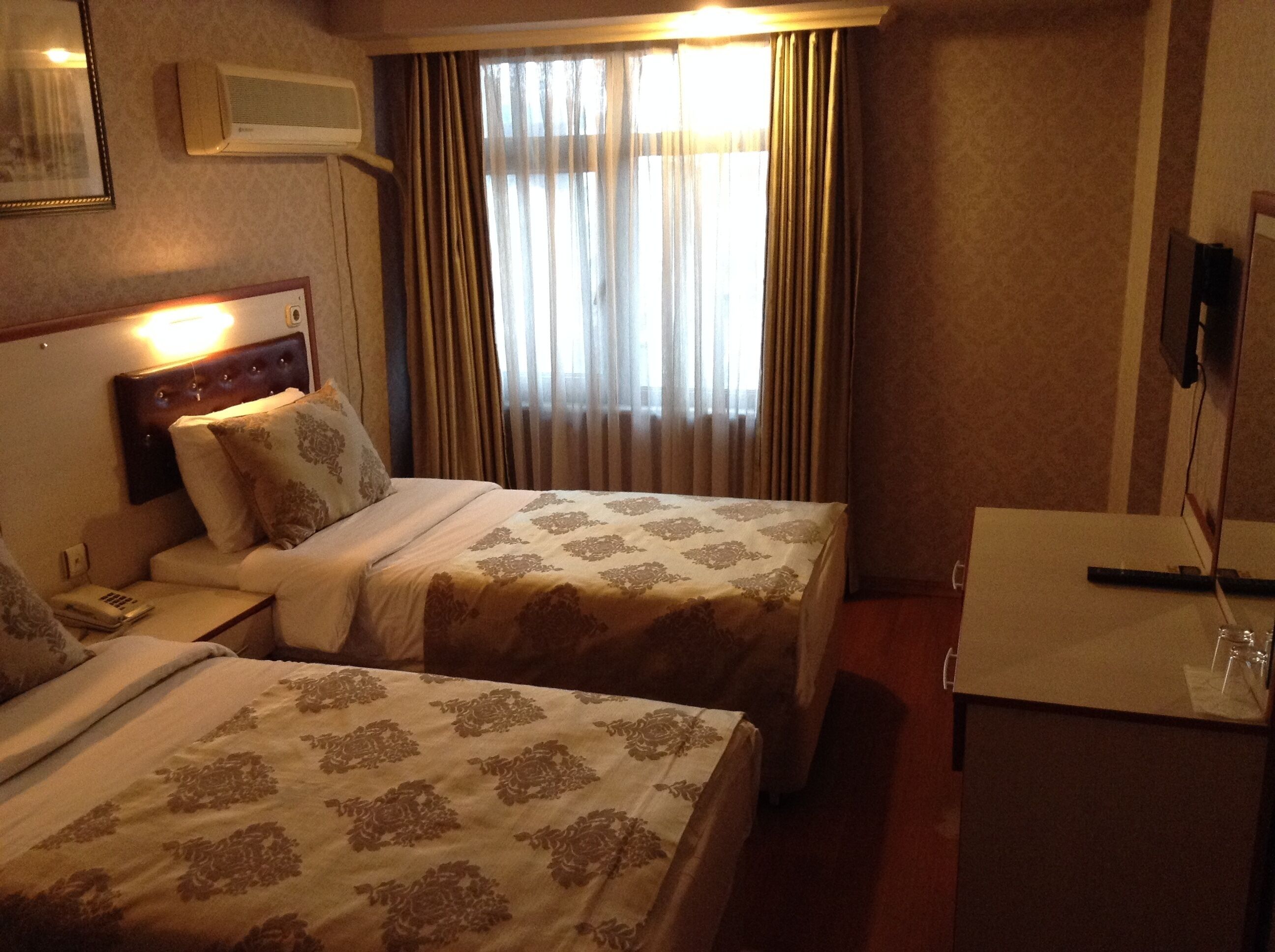 A hotel room with two beds, a bedside table with a lamp, a TV, and a window with curtains.