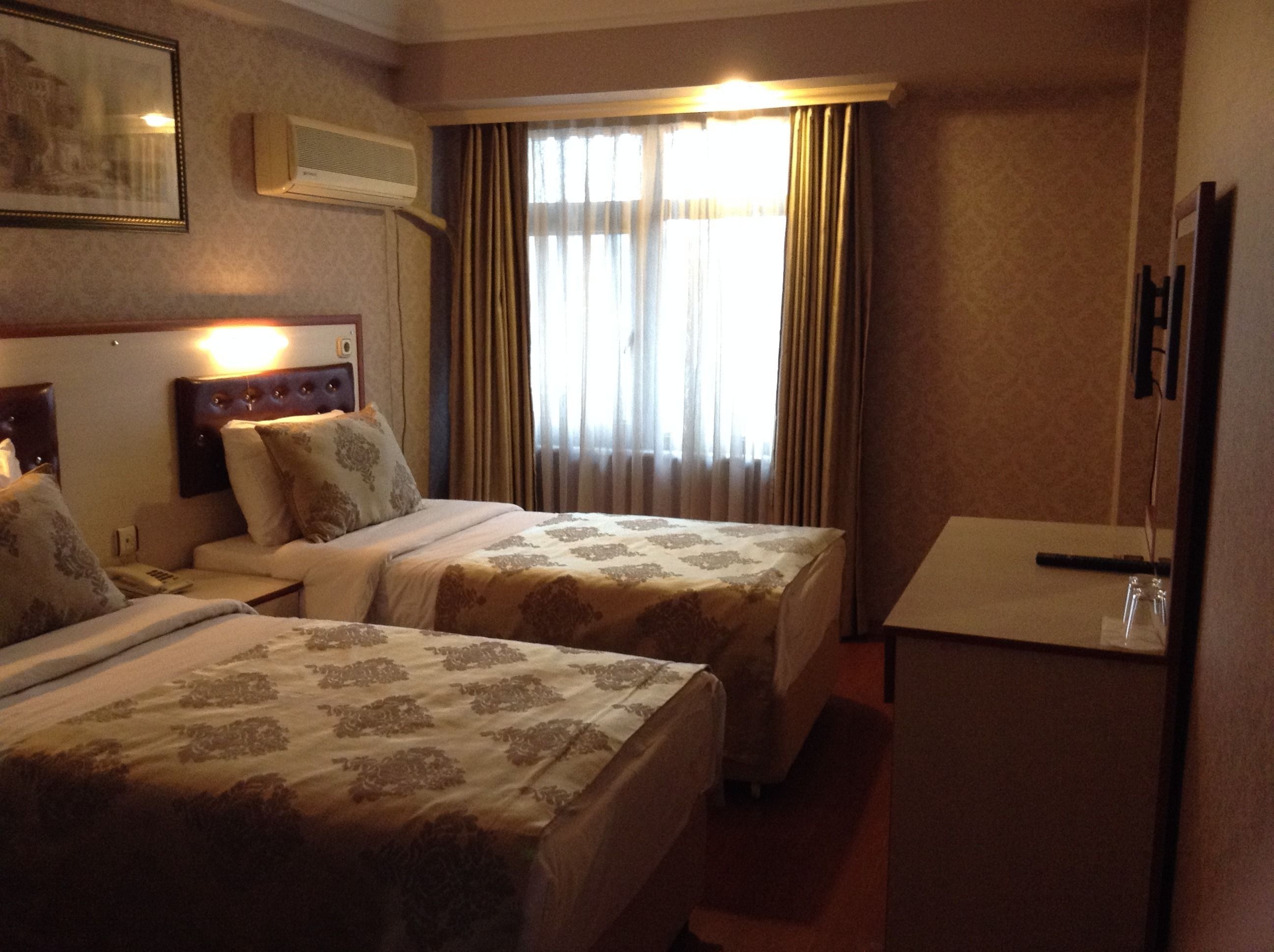 A hotel room with two beds, a bedside table, a lamp, an air conditioning unit, and a window with curtains.