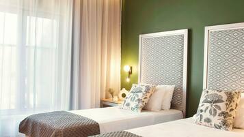 A hotel room with two beds, a large window with sheer curtains, a green accent wall, and a nightstand with a lamp.