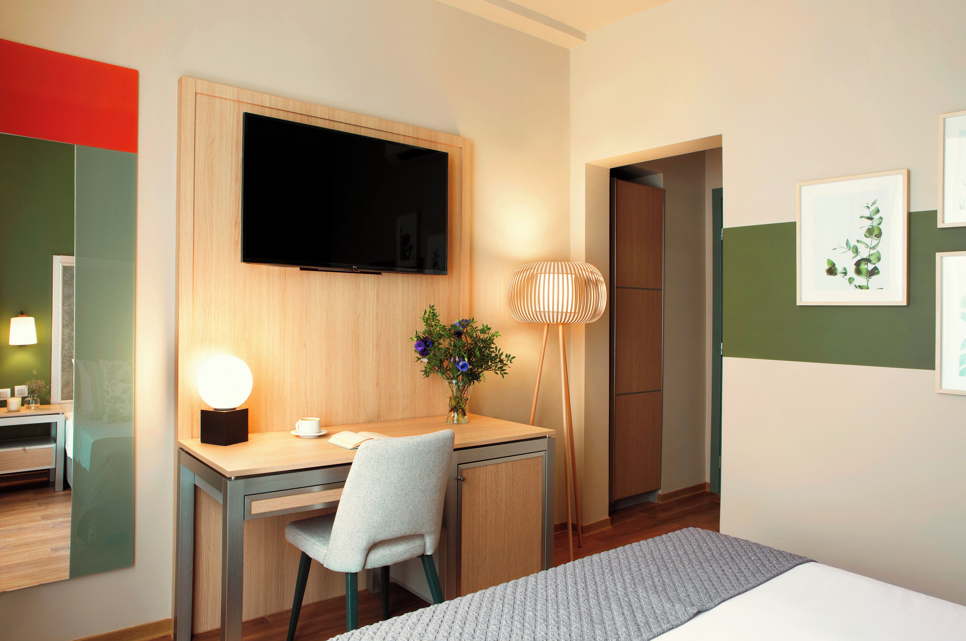 A hotel room with a bed, a desk with a chair, a flat-screen TV mounted on the wall, a floor lamp, and framed artwork on the wall.