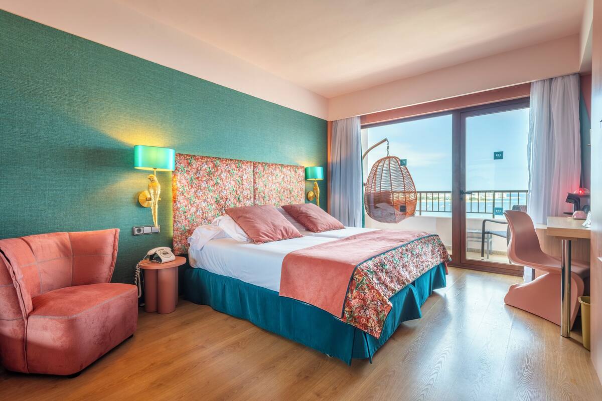 Habitación de hotel con cama, escritorio, silla, mecedora y vistas al exterior.