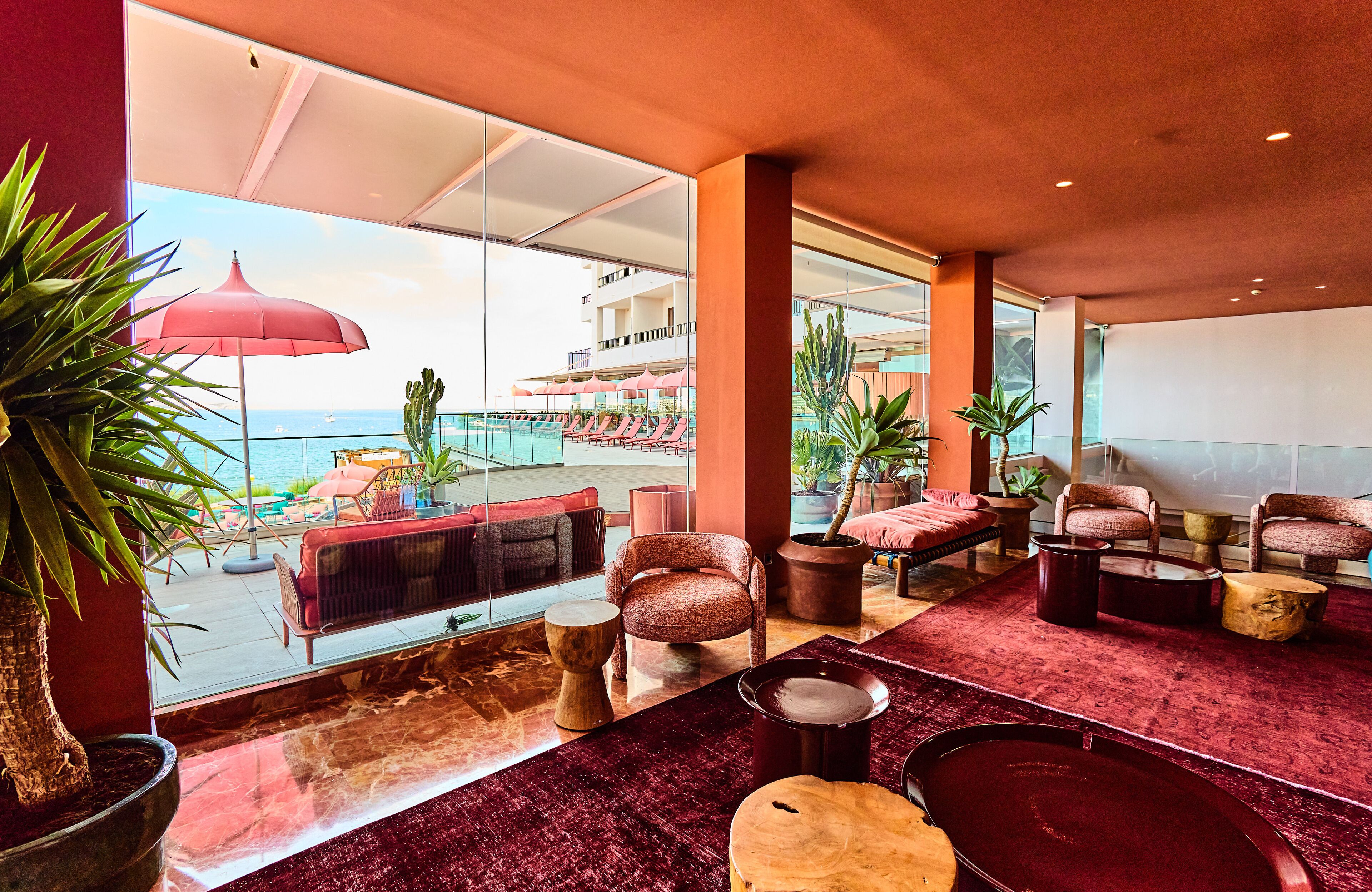 A modern lounge area with a view of the sea, featuring comfortable seating, a red carpet, and potted plants.