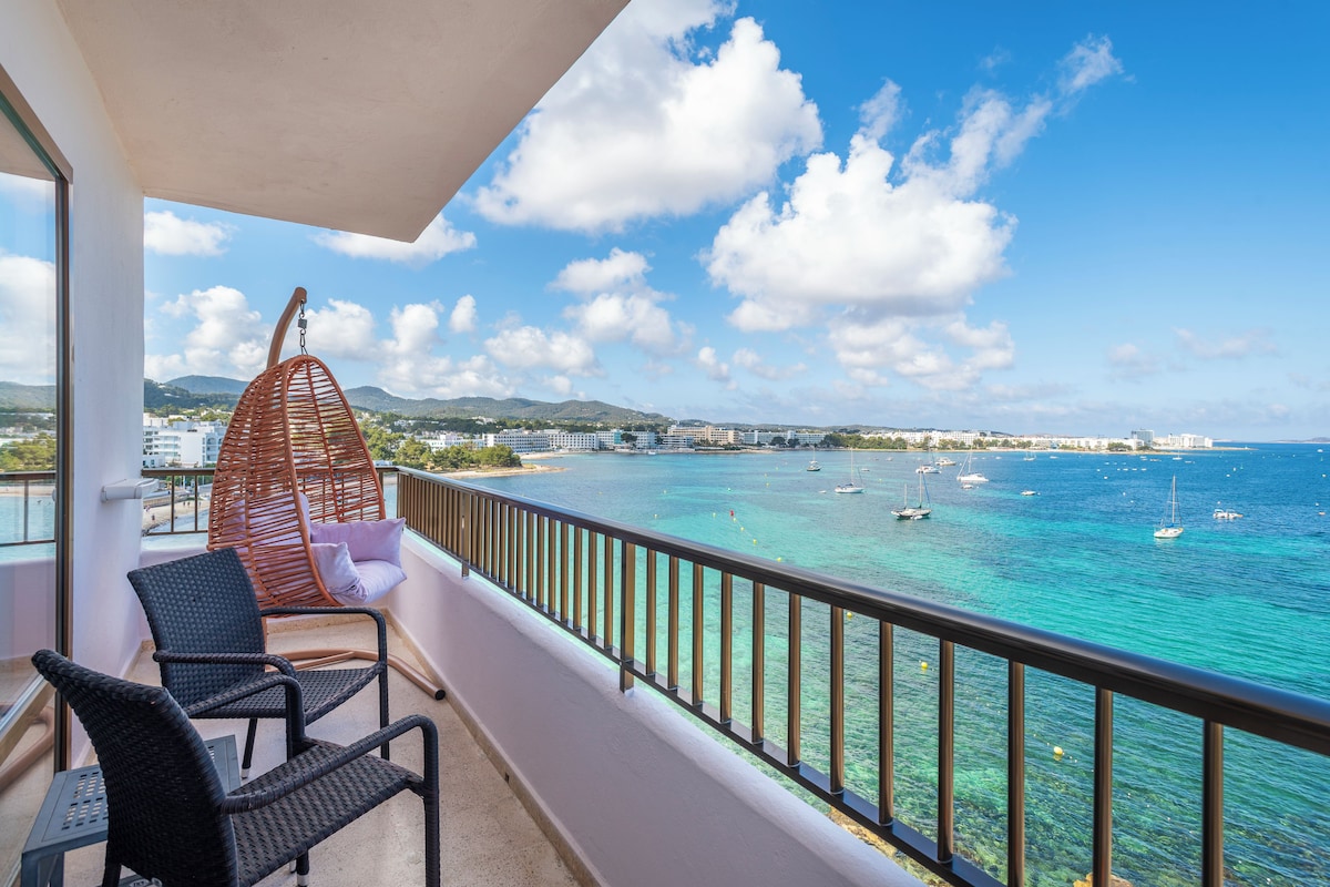 A balcony with wicker chairs, a hanging chair, and a view of a coastal area with boats.