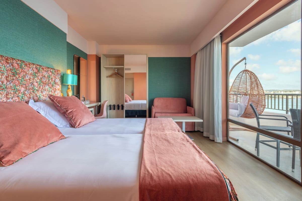 Una habitación de hotel con dos camas, una zona de estar con una silla y una mesa, un ventanal con vistas y un espejo.