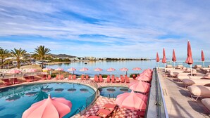 Un espace aménagé au bord de la piscine, avec plusieurs chaises longues et des parasols roses, un ciel dégagé et une étendue d'eau en arrière-plan.