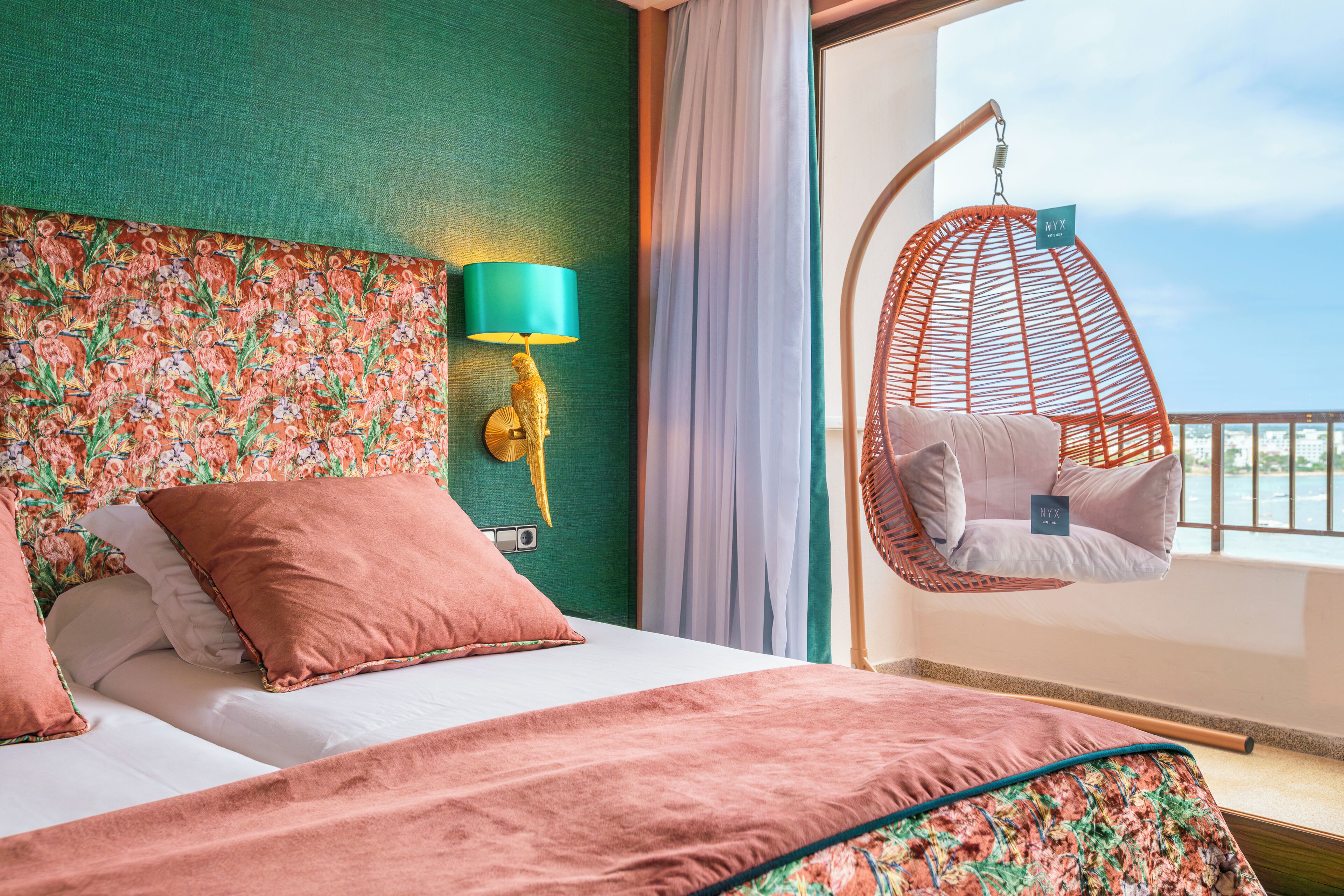 Habitación de hotel con cama, una silla colgante, lámpara de pared y vistas al exterior.