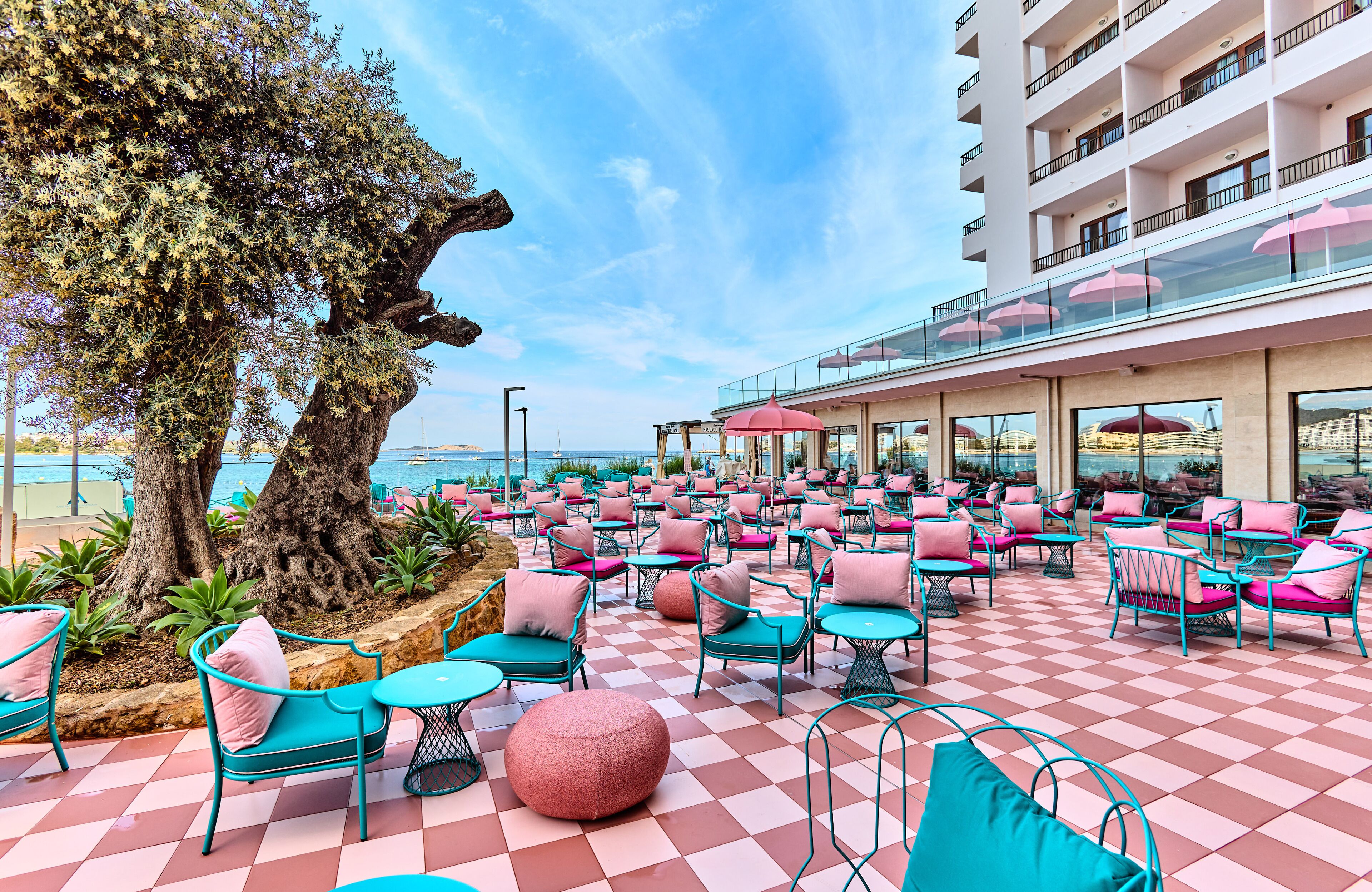 Eine Terrasse mit Schachbrettböden, türkisfarbenen und rosafarbenen Stühlen und einem großen Baum.