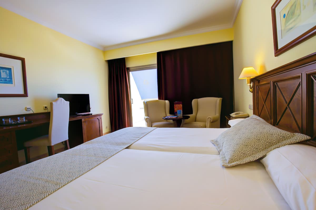 A hotel room with two beds, a desk with a computer, a chair, a lamp, and a window with curtains.
