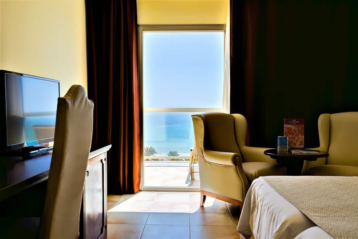 A hotel room with a bed, two armchairs, a small table, a TV, and a large window with a view of the ocean.