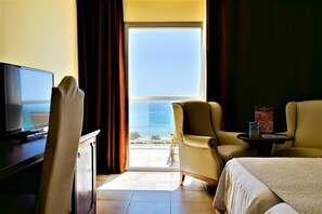 Een hotelkamer met een bed, twee fauteuils, een kleine tafel, een televisie en een groot raam met uitzicht op de oceaan.