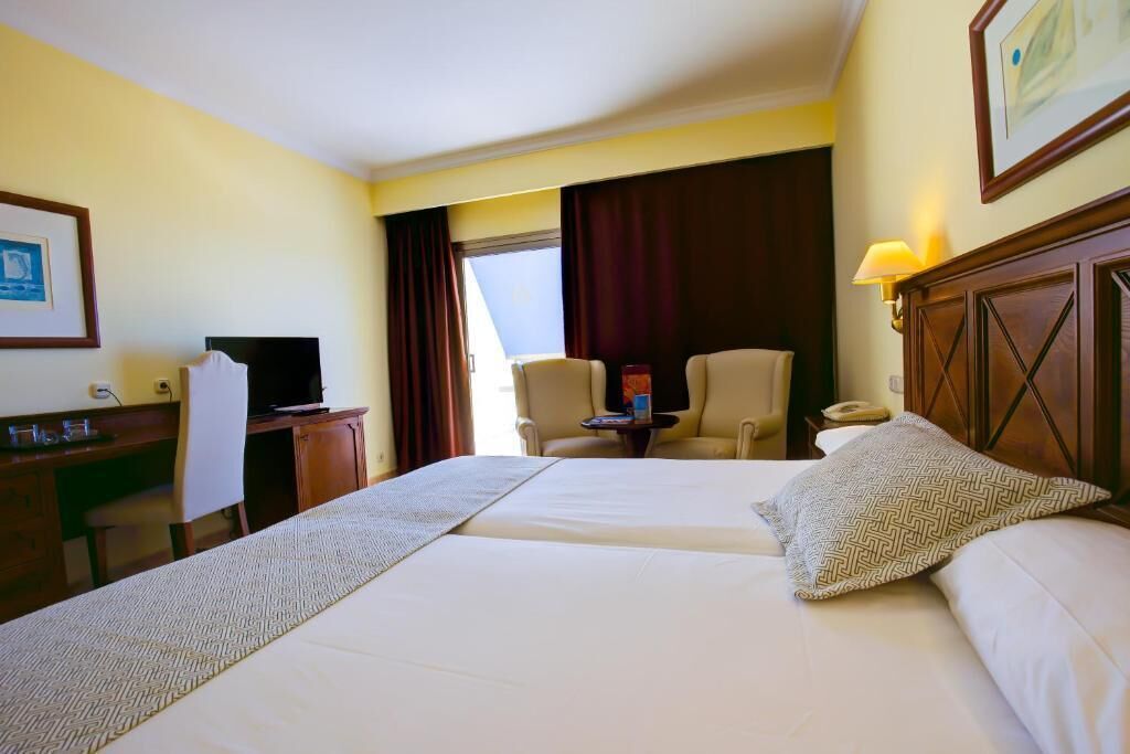 A hotel room with two beds, a desk, a chair, a television, and a window with curtains.