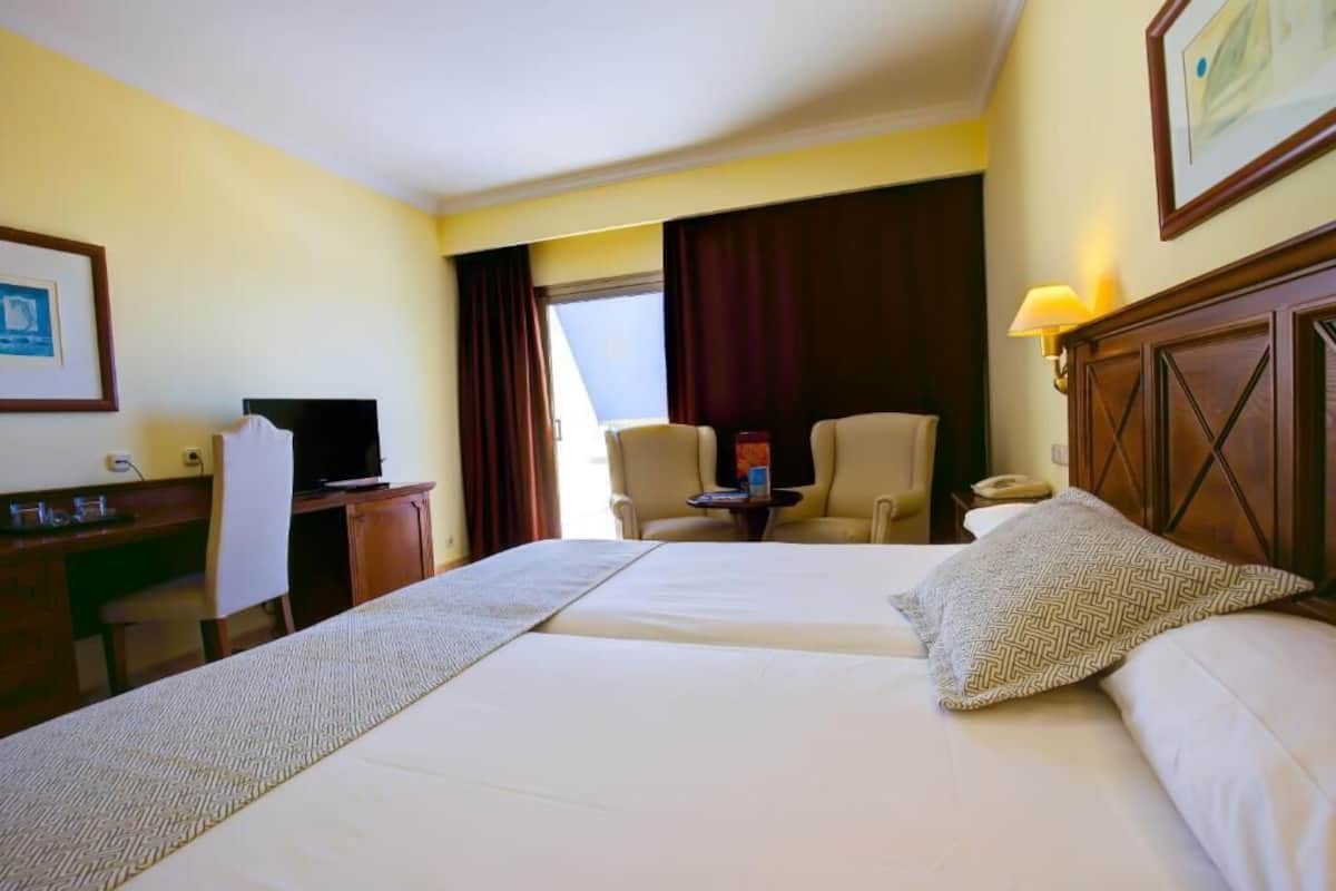 A hotel room with two beds, a desk, a chair, a television, and a window with curtains.