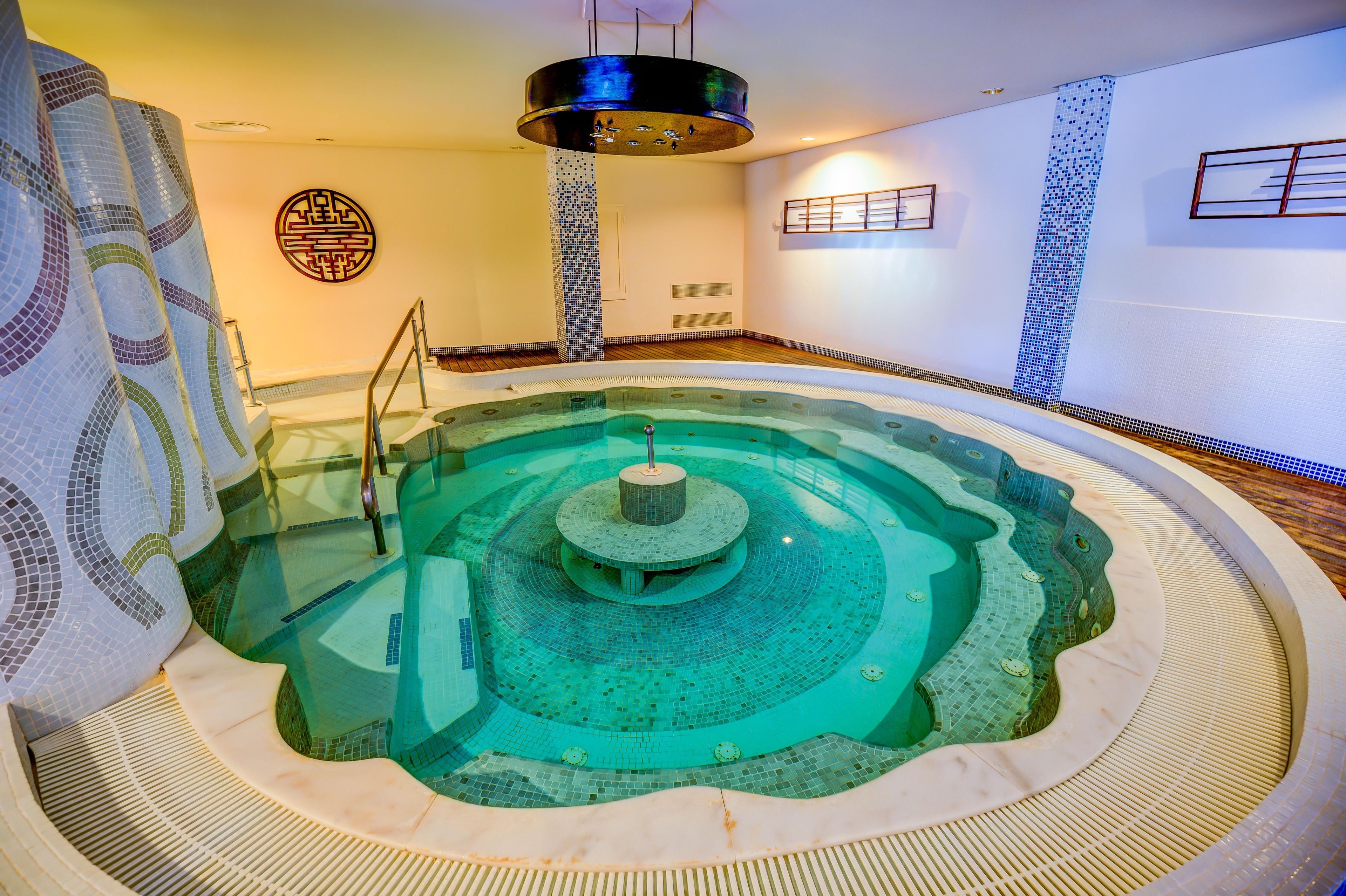 Una amplia piscina interior con una fuente central, rodeada por un solárium con baldosas y una pared de mosaicos.