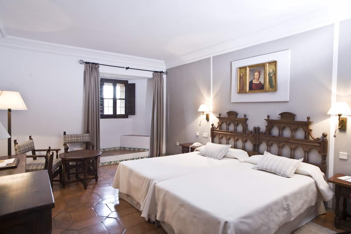 Ein Hotelzimmer mit zwei Betten, einem Esstisch, Stühlen, einem kleinen Tisch, einer Lampe, einem Gemälde und einem Fenster mit Vorhängen.