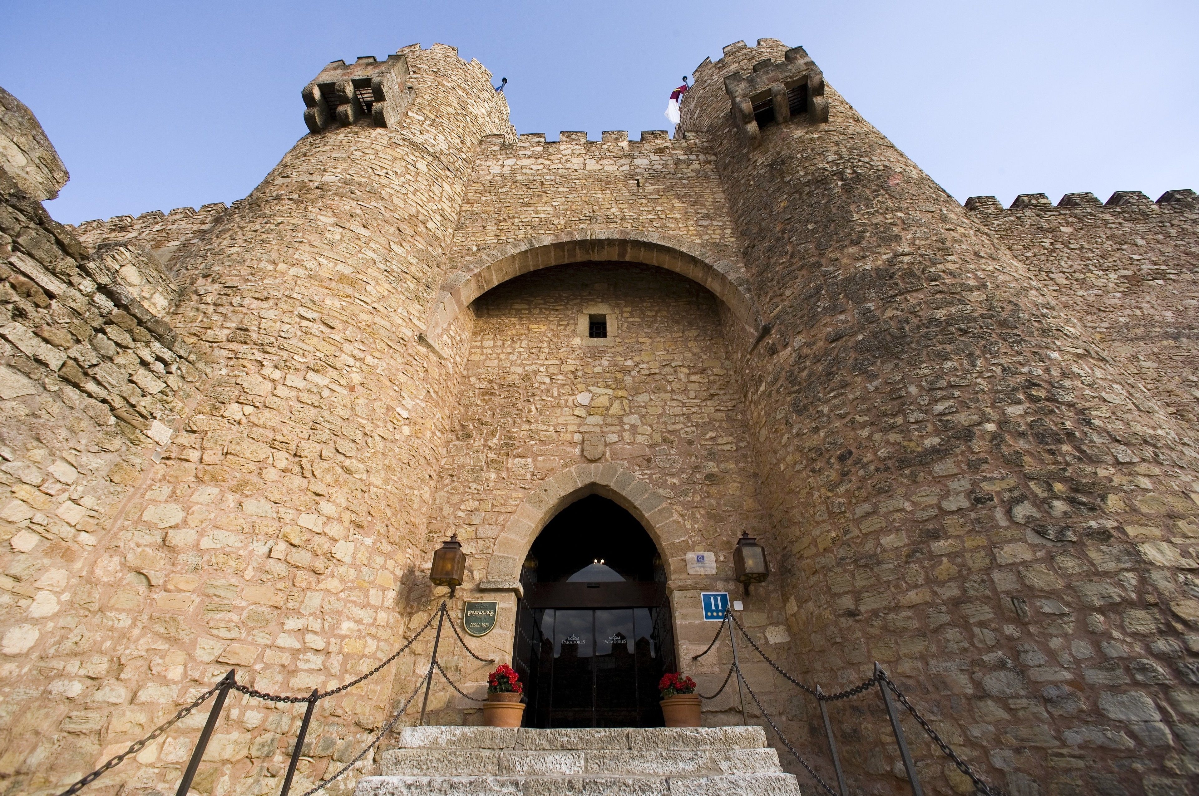 A stone castle entrance with a central archway, two lanterns on either side, and a sign indicating a historical site.
