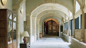Un pasillo con arcos de piedra, un aparador de madera con puertas de cristal, una mesa con una lámpara y una puerta al final.
