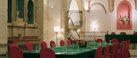 Un salón de banquetes con una larga mesa dispuesta para un evento formal, rodeado de sillas rojas, un candelabro y techos abovedados.
