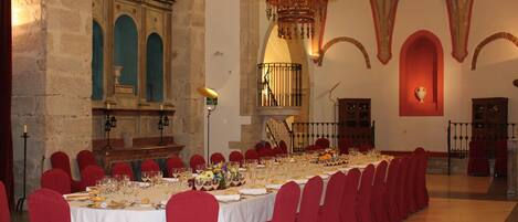 Un salón de banquetes con largas mesas dispuestas para un evento formal, sillas rojas y una araña.