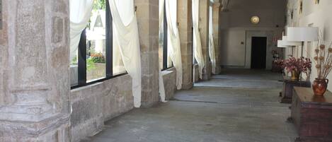 A corridor with stone columns, large windows with white drapes, and a wooden bench with a vase.
