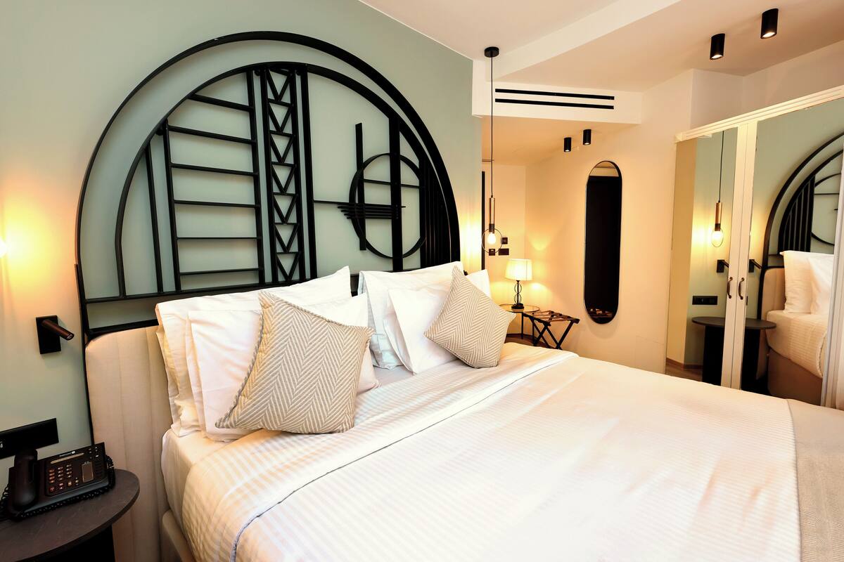 Una cama bien tendida con sábanas blancas y almohadas estampadas, un teléfono en la mesita de noche y un espejo colgado en la pared.