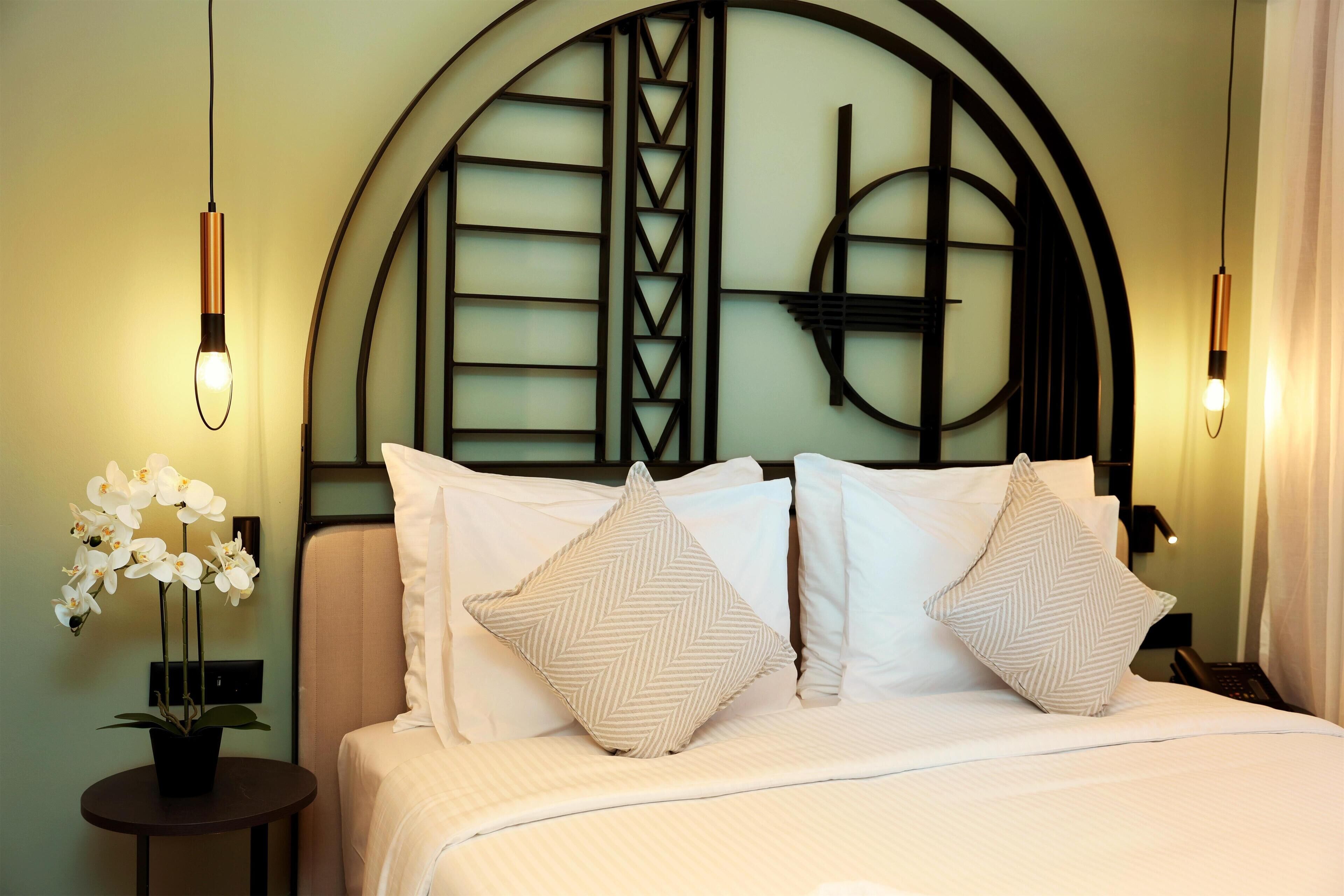 Een net opgemaakt bed met twee kussens, een klein tafeltje met een bloeiende orchidee en een muur met een decoratief metalen ontwerp.