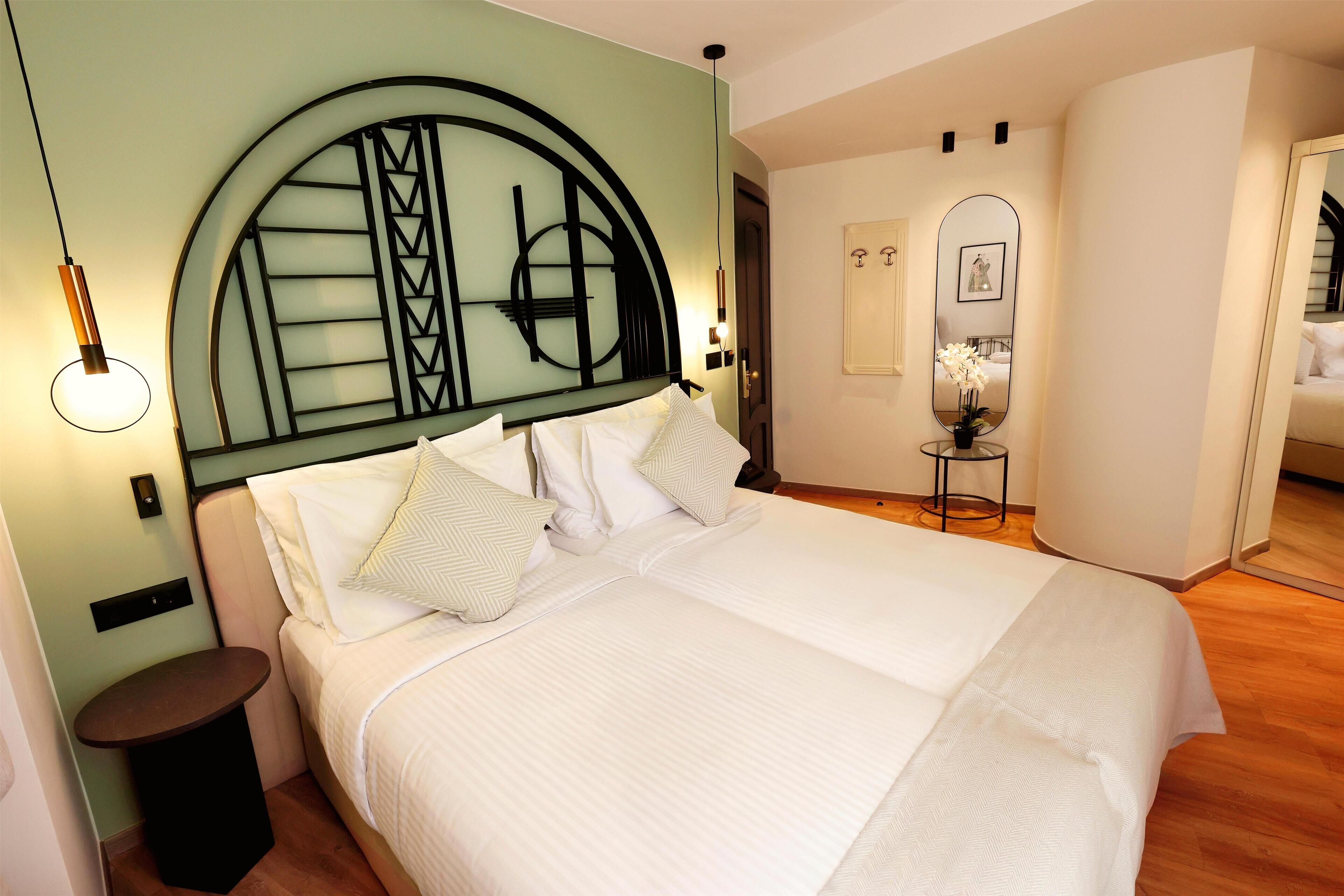 Een net opgemaakt bed met witte lakens, een rond nachtkastje en een spiegel aan de muur.