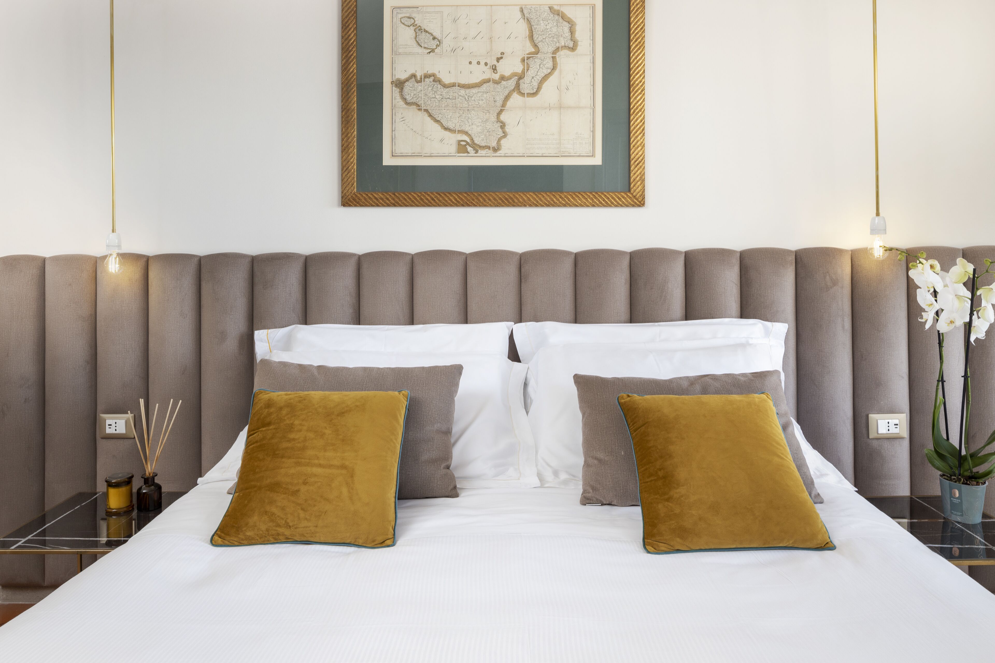 Una cama bien tendida con dos almohadas, un mapa enmarcado en la pared y una mesita de noche con un florero y difusor de cañas.