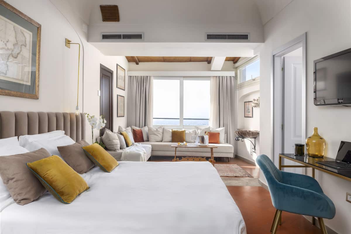 A hotel room with a large bed, a sofa, a desk with a laptop, and a TV on the wall.