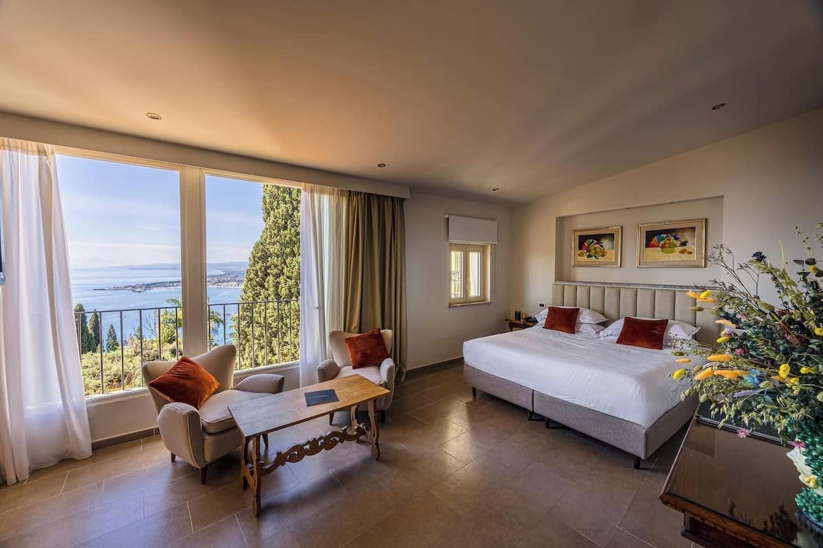 A spacious bedroom with a large bed, a sitting area with a table, and a view of the sea.
