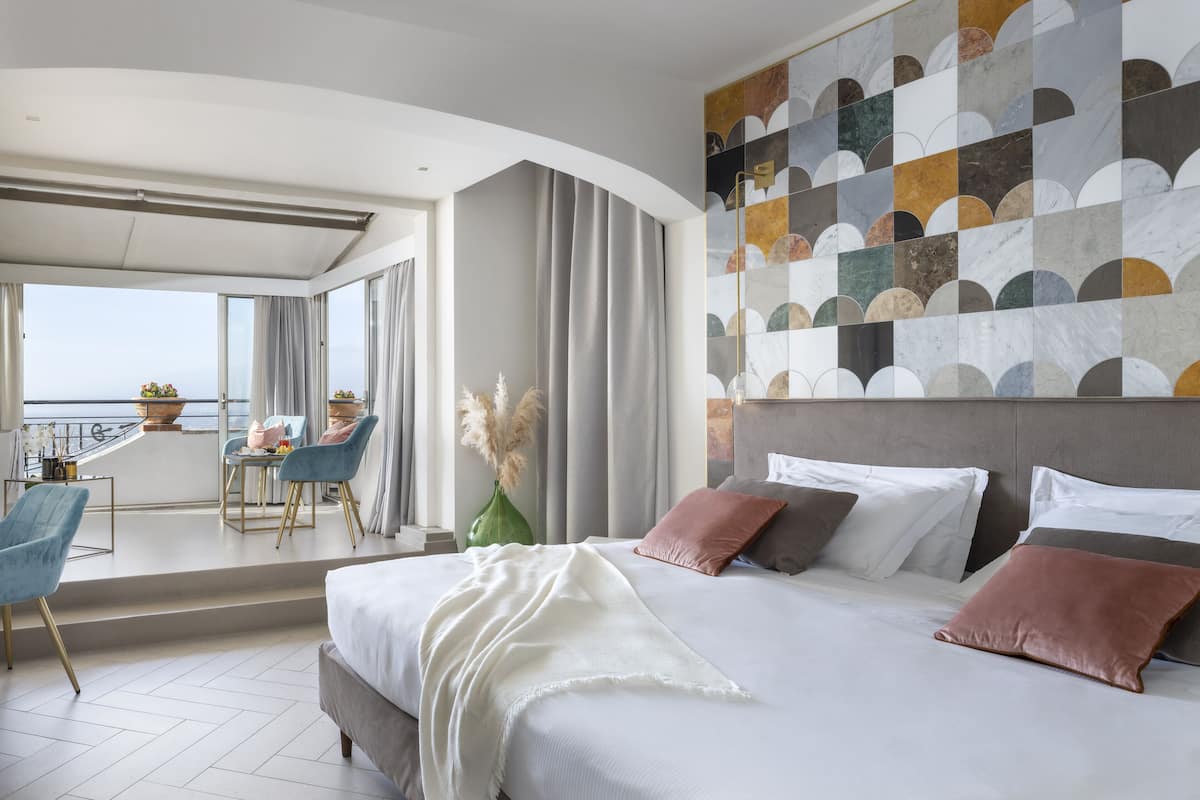 A modern hotel room with a large bed, a balcony with a table and chairs, and a view of the sea.