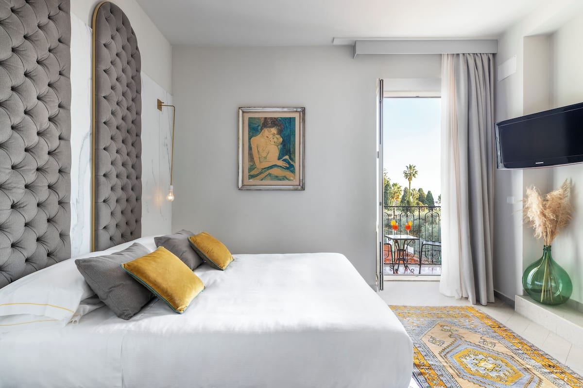 A hotel room with a large bed, a painting on the wall, a TV, and a balcony with a view.