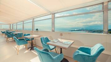 Un comedor moderno con sillas y mesas color turquesa, amplias ventanas que ofrecen vistas al mar y al paisaje urbano.