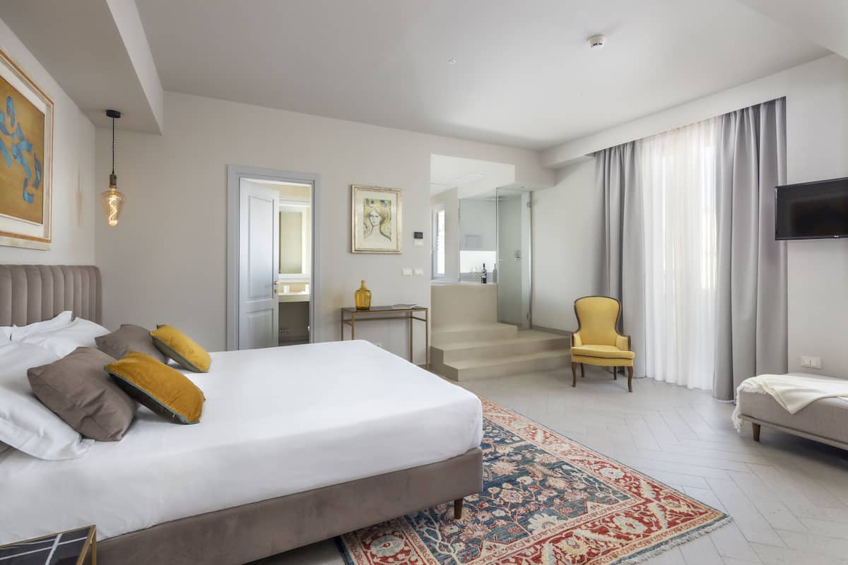 A modern hotel room with a large bed, a sitting area with a chair, a TV, and a bathroom with a glass shower.