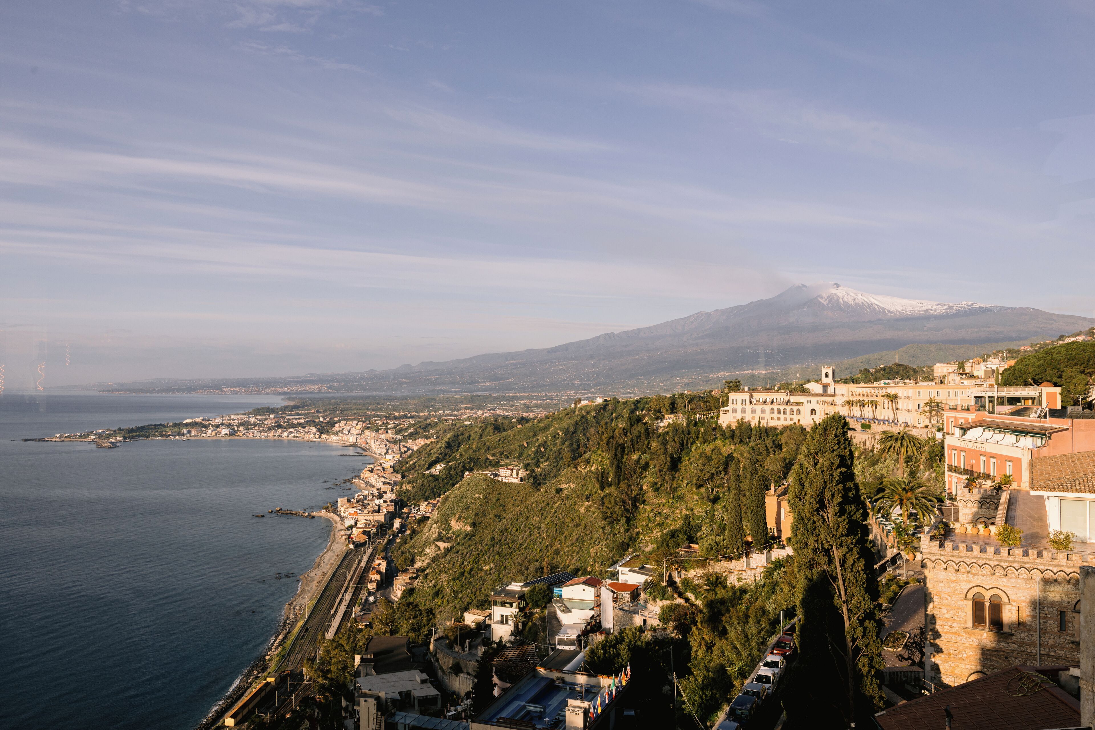 A coastal town with a prominent mountain in the background, a mix of residential and historical buildings, and a clear view of the sea.