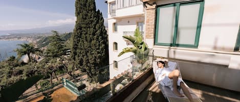 A balcony with a view of a coastal landscape, a tall cypress tree, and a sign for "TACOMA SPA".