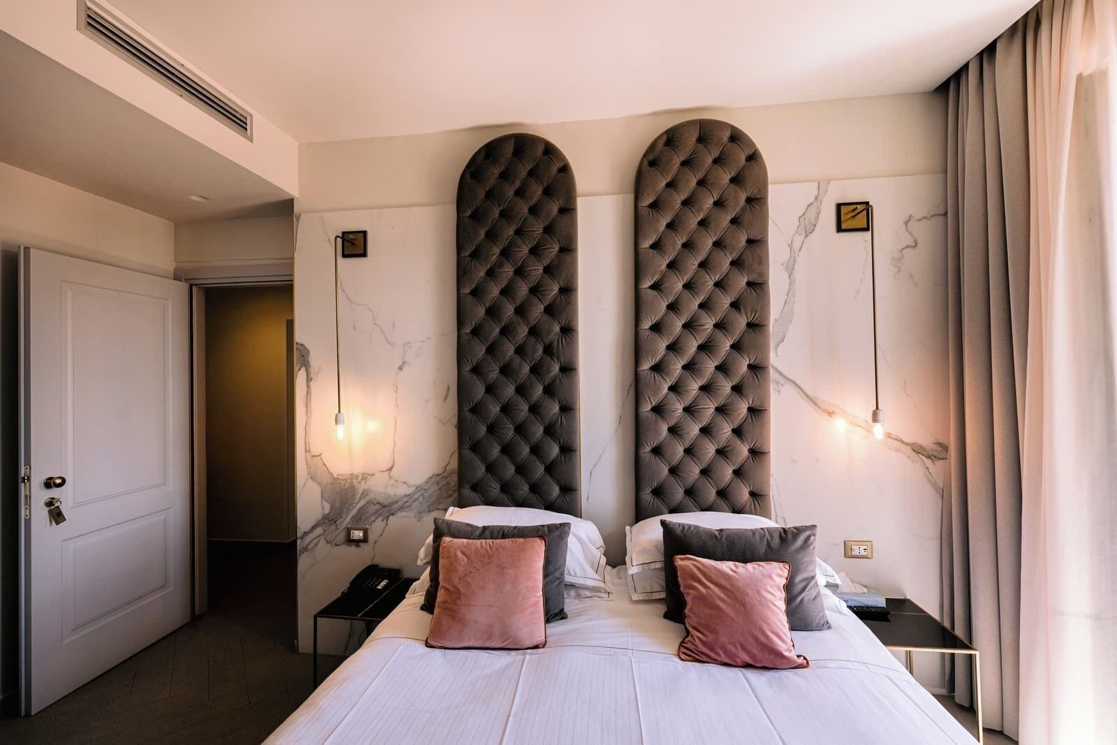 Ein modernes Schlafzimmer mit einem großen Bett, zwei gepolsterten Kopfteilen, Marmorwänden und einem Nachttisch mit Lampe.
