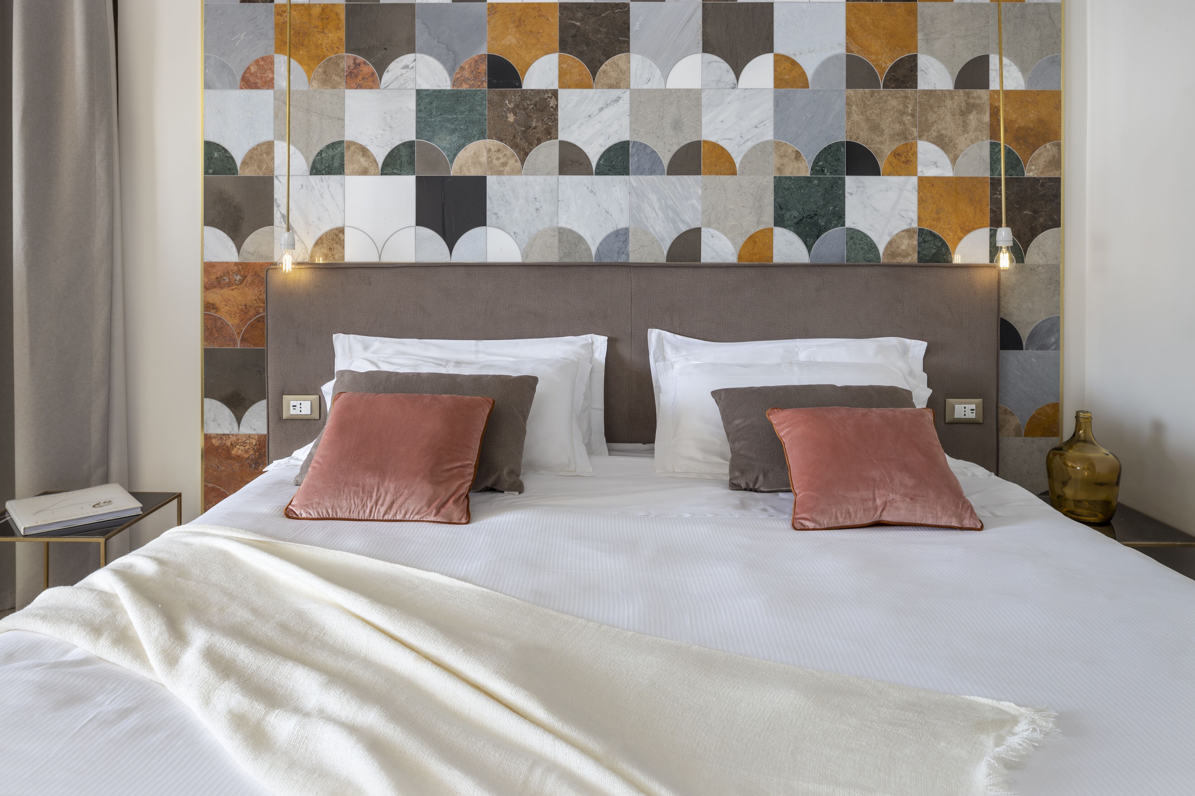 Una cama bien hecha con sábanas blancas y dos almohadas, un papel tapiz estampado en la pared de fondo y una mesita de noche con una lámpara.