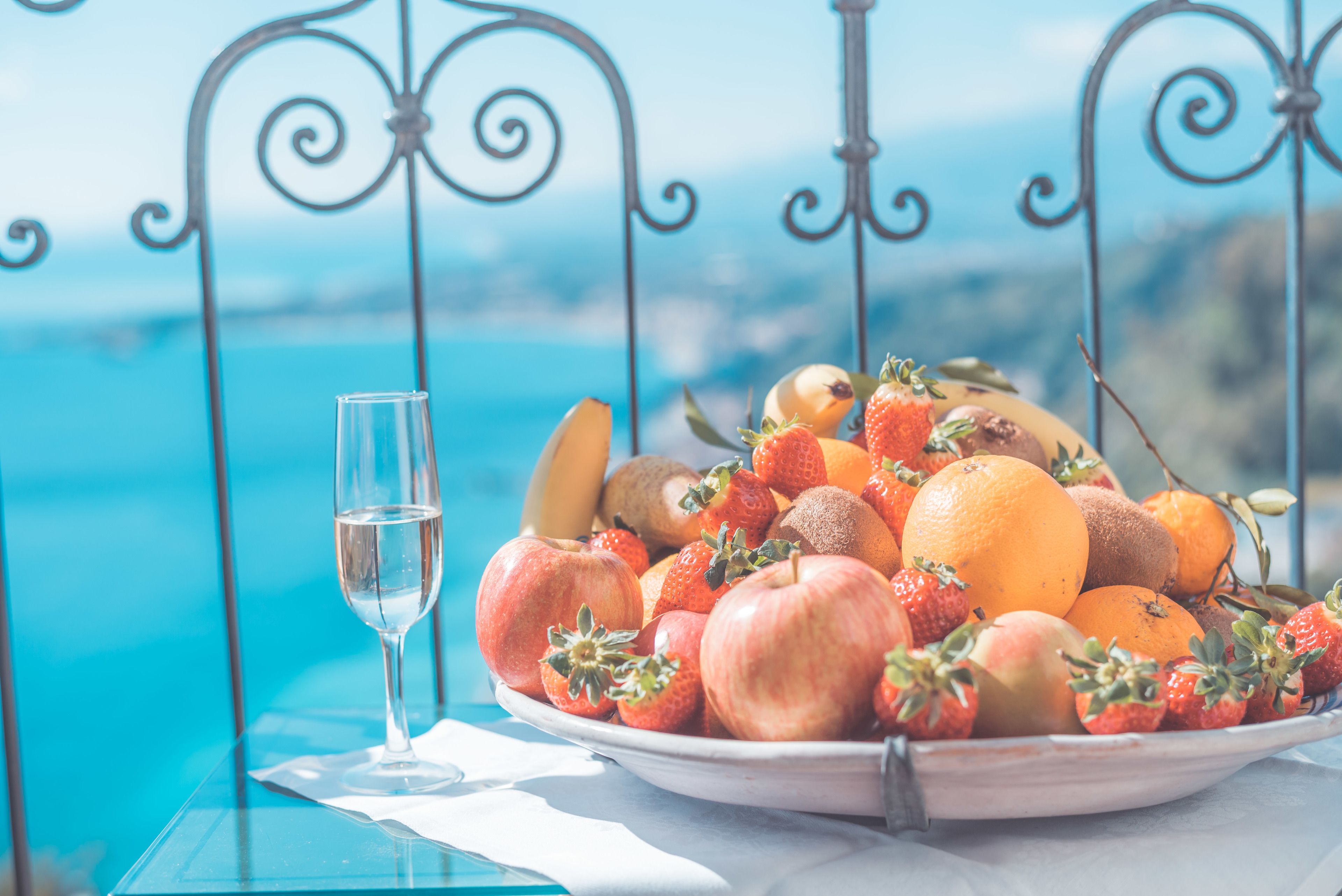 A table with a fruit bowl and a glass of water, set against a scenic view.