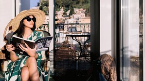 A person sitting on a chair by a window with a view of a cityscape and a balcony with wicker furniture. - Hotel Villa Paradiso (Taormina)