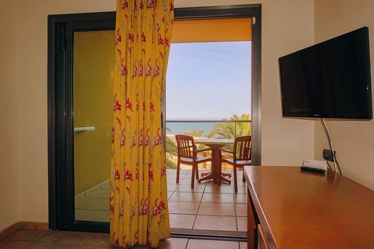 A room with a sliding glass door leading to a balcony with a table and chairs, a flat-screen TV mounted on the wall, and a power outlet on the table.