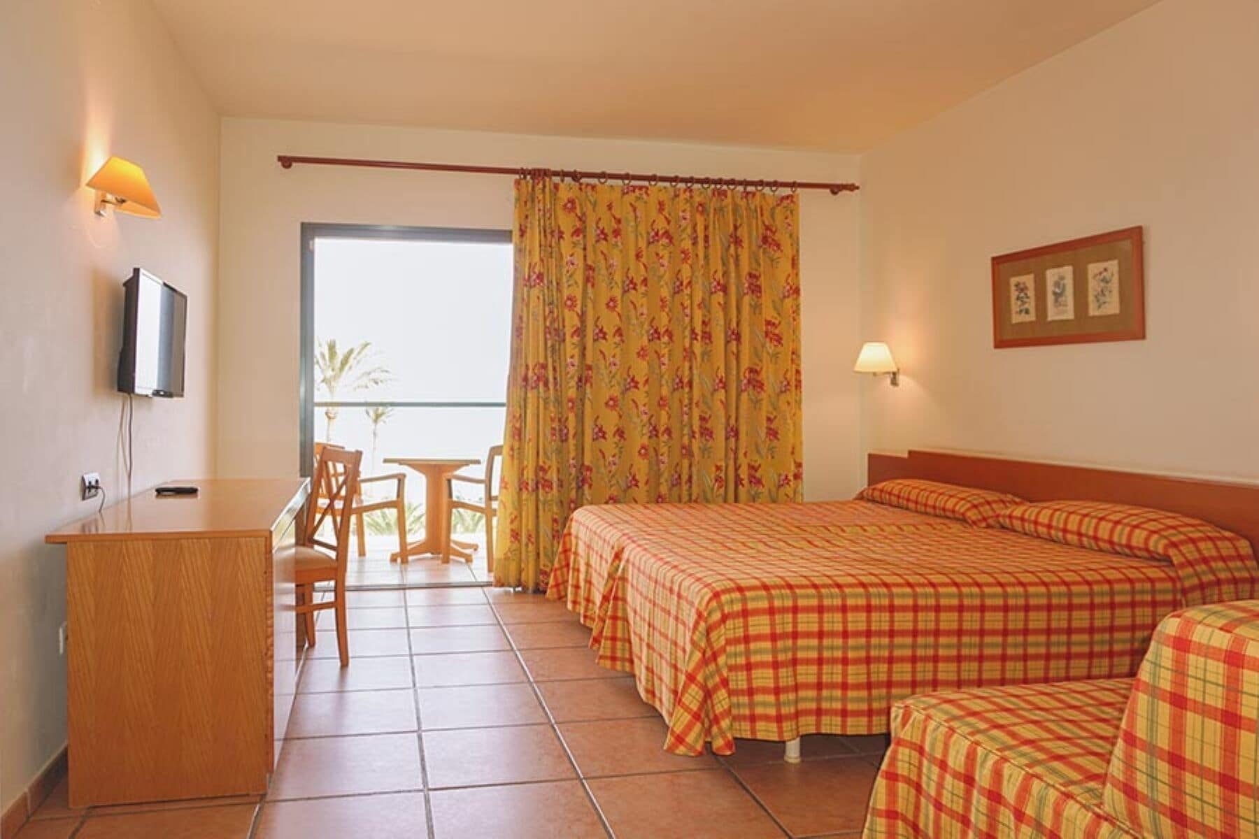 Une chambre d’hôtel comprenant un lit, un bureau, une chaise, une télévision, une fenêtre avec des rideaux et une lampe fixée au mur.