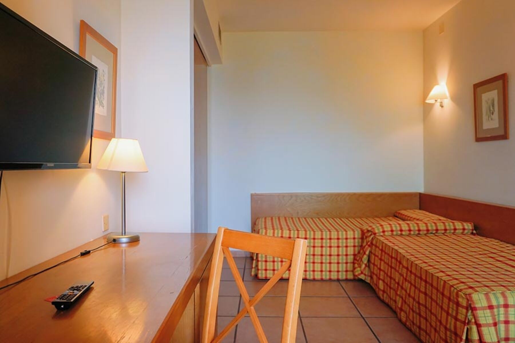 A hotel room with a television, a lamp, a wooden chair, and two beds with checkered bedding.