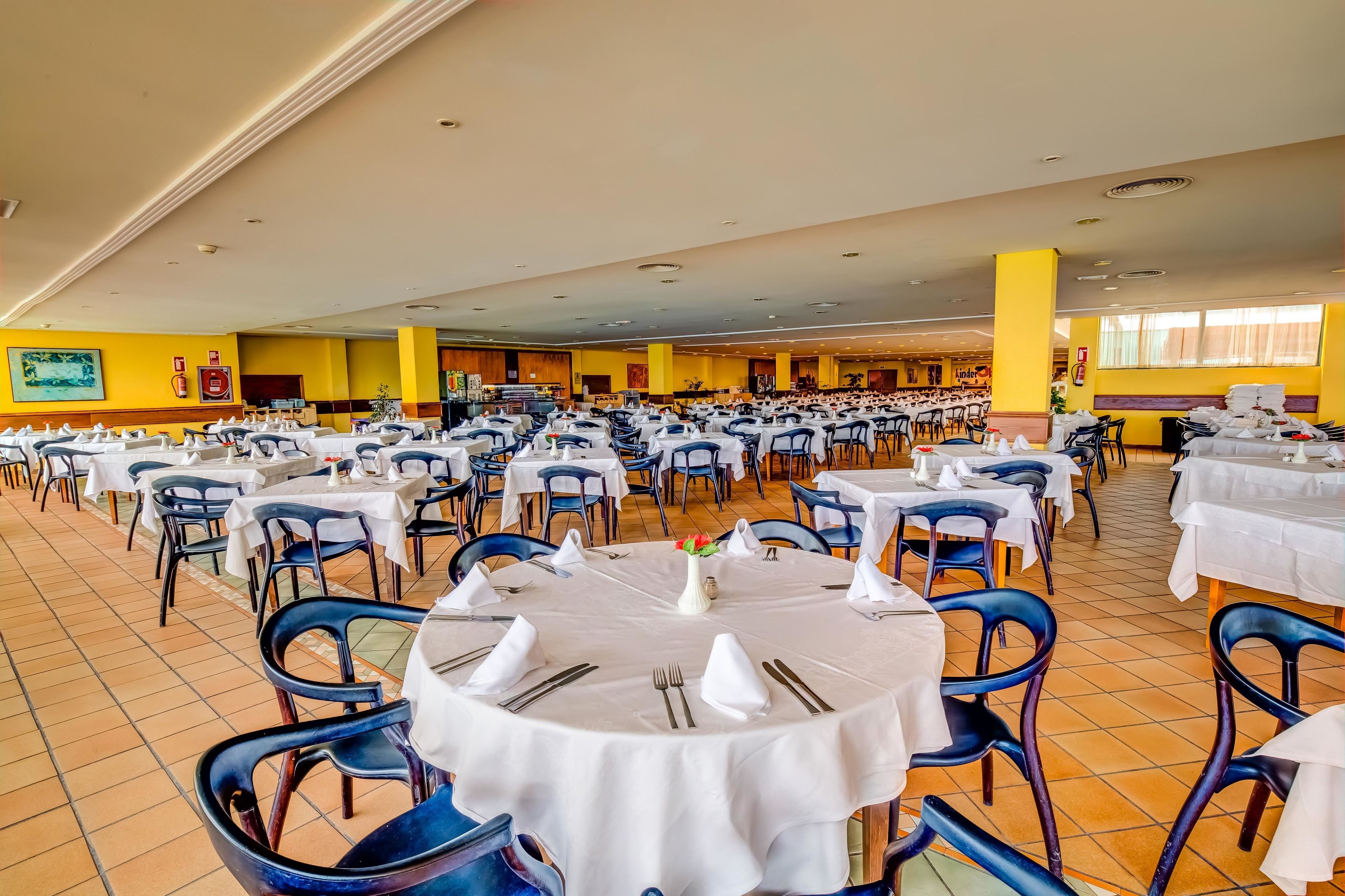 Um amplo salão de banquetes com várias mesas redondas preparadas para refeições, com toalhas brancas e cadeiras azuis.