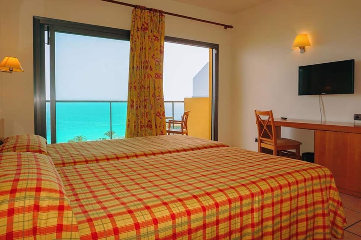 A hotel room with a bed, a desk, a chair, a TV, and a balcony with a view of the ocean.