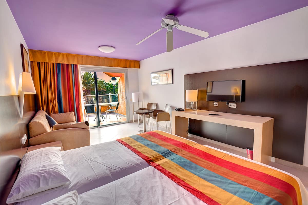 A hotel room with a bed, a desk, a TV, and a balcony with orange curtains.