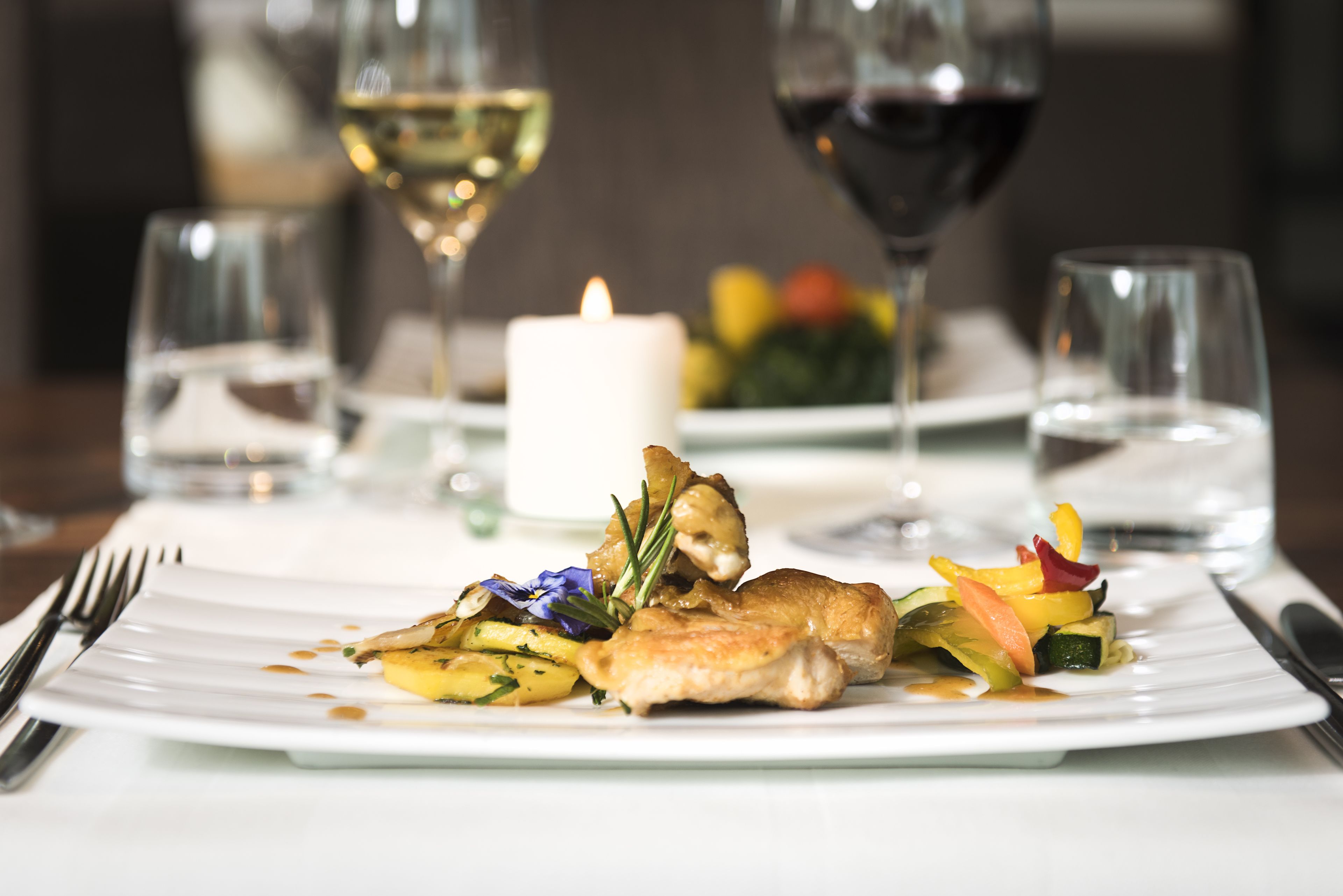 A white plate with a chicken dish, vegetables, and a candle on a table.