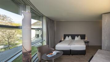 A modern hotel room with a large window, a bed with two pillows, a bedside table with a magazine, and a chair.