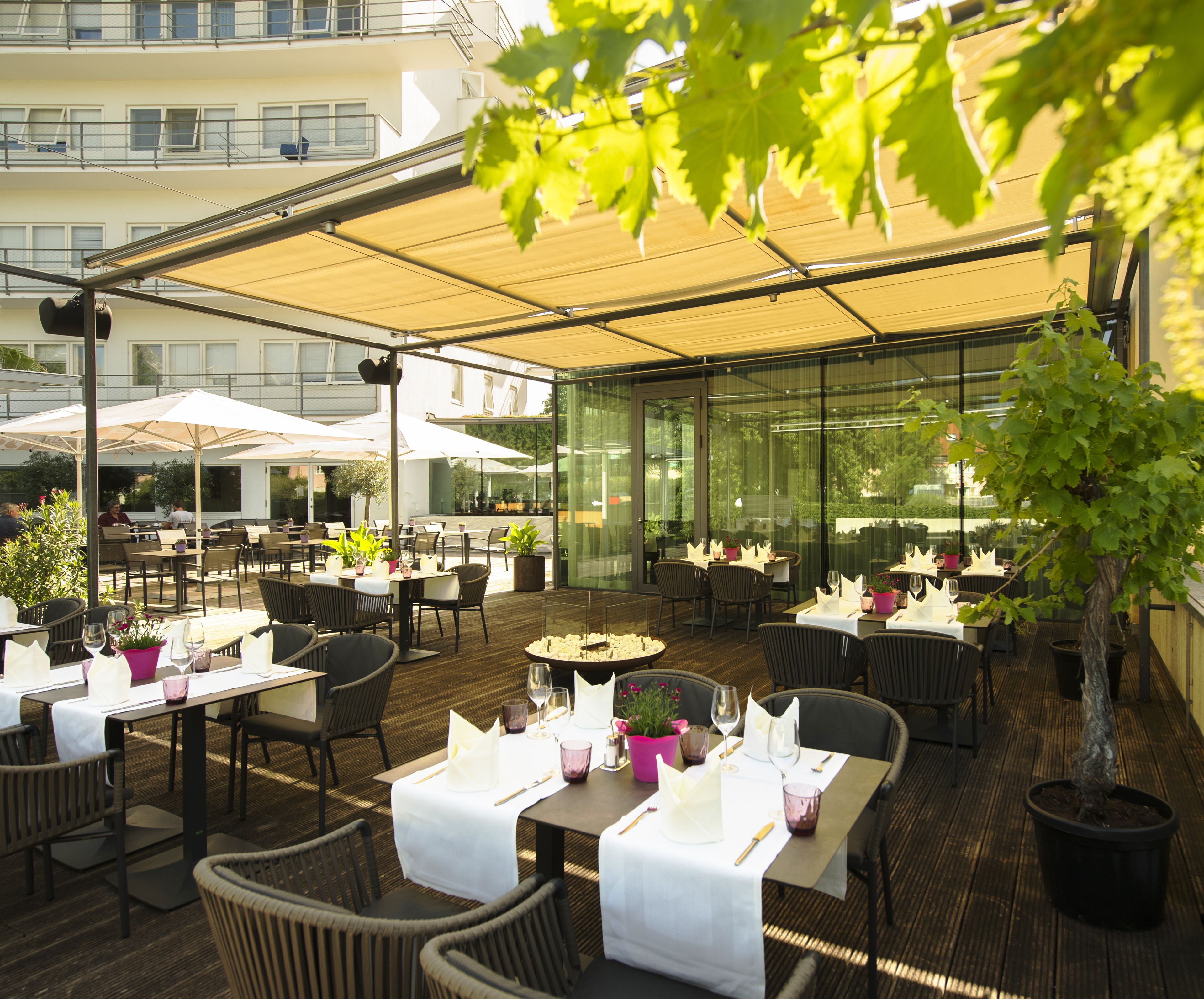 An outdoor dining area with tables and chairs, covered by a canopy, and surrounded by potted plants.